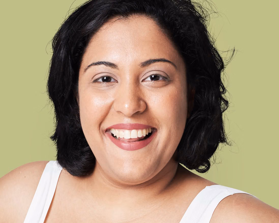 A woman with short black hair and a brown shirt smiles broadly while looking toward the camera against a white background.