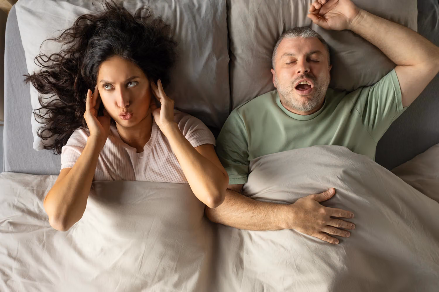 A man snores in bed while his partner covers her ears with her fingers.