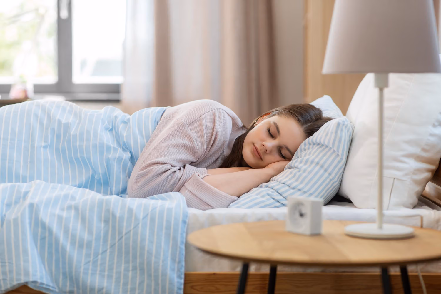 A woman sleeps on her side in a bed with blue and white striped bedding, next to a bedside table with a lamp and a small clock.