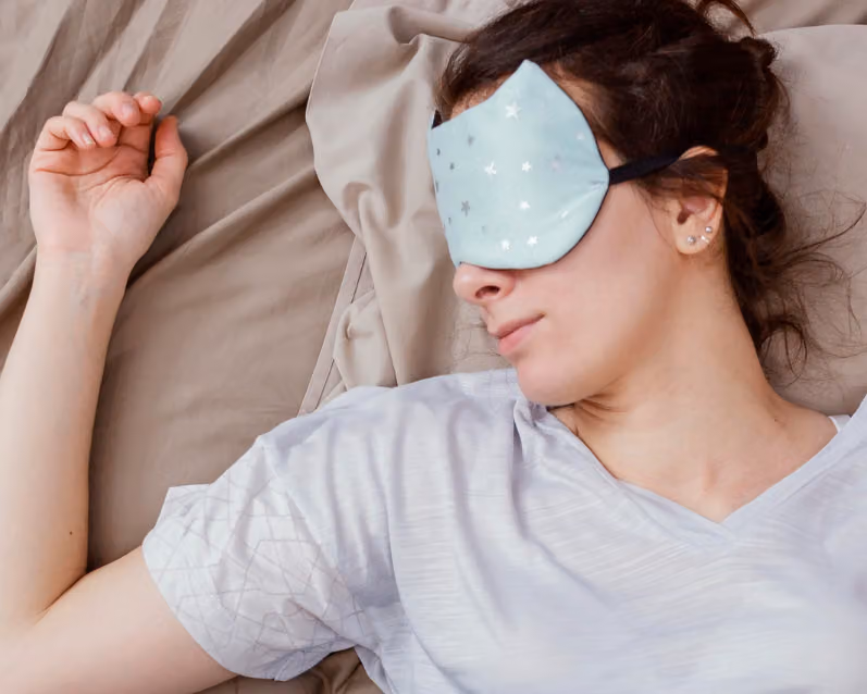 A woman wearing a light blue eye mask with stars lies on a bed, resting on her side with one arm bent above her head.