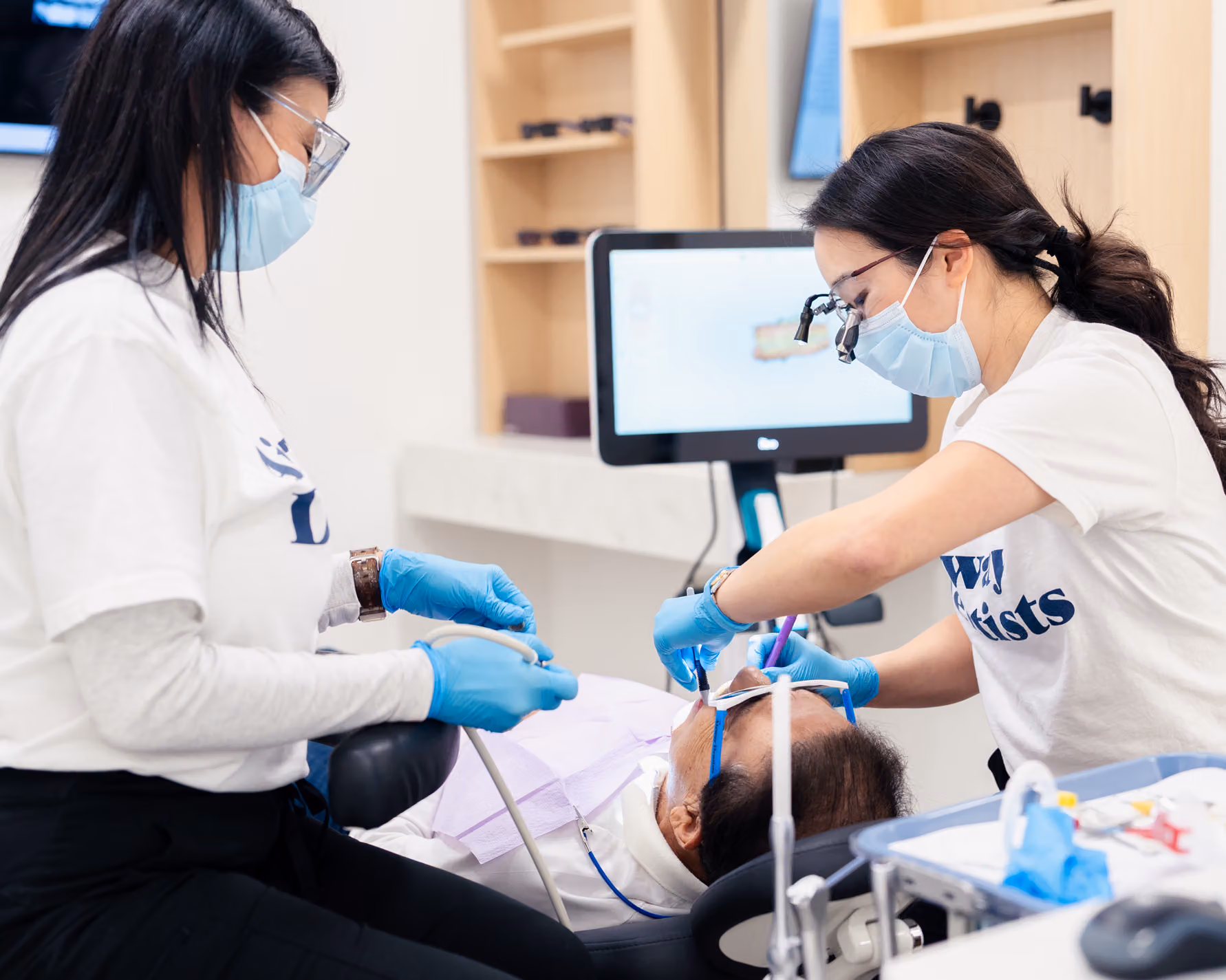 Two dental professionals in masks and gloves perform a dental procedure on a patient in a modern clinic setting with equipment visible.