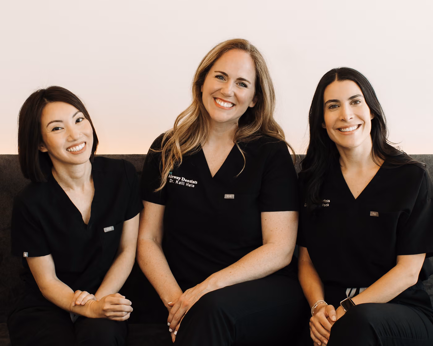 Three members of The Airway Dentists sitting and smiling wearing matching black "Airway Dentists" scrubs.
