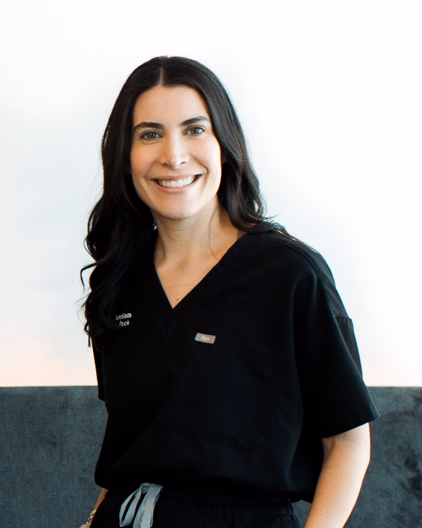 Woman with long dark hair wearing black medical scrubs smiles while standing against a light background.