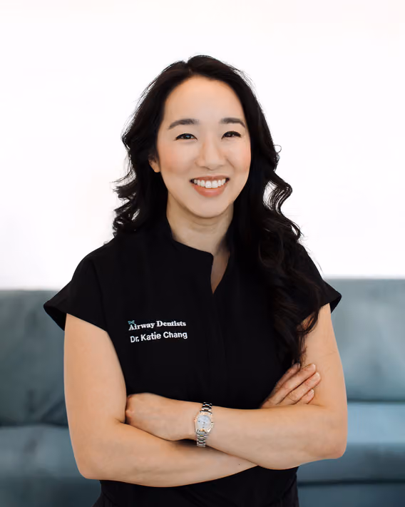 A woman with long dark hair, wearing a black top with "Dr. Katie Chang" embroidered, stands with arms crossed and smiles at the camera.