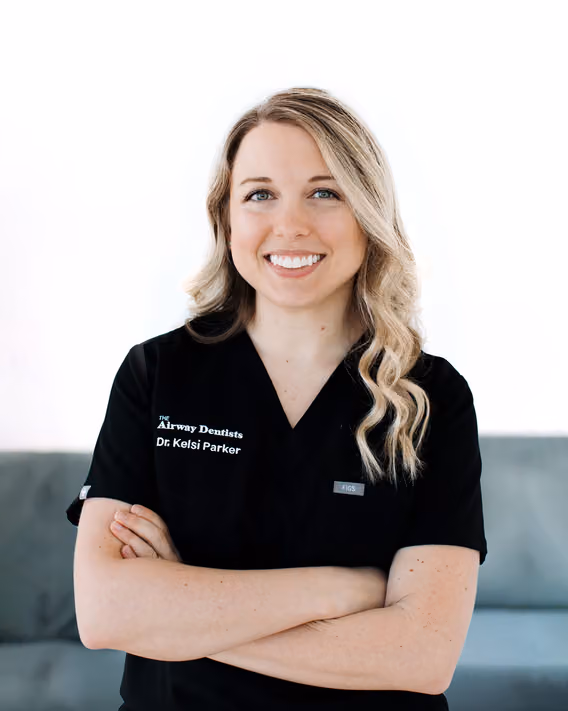 A woman with long blonde hair in black scrubs stands with arms crossed, smiling at the camera. Her uniform reads “The Airway Dentists Dr. Kelsi Parker.”.