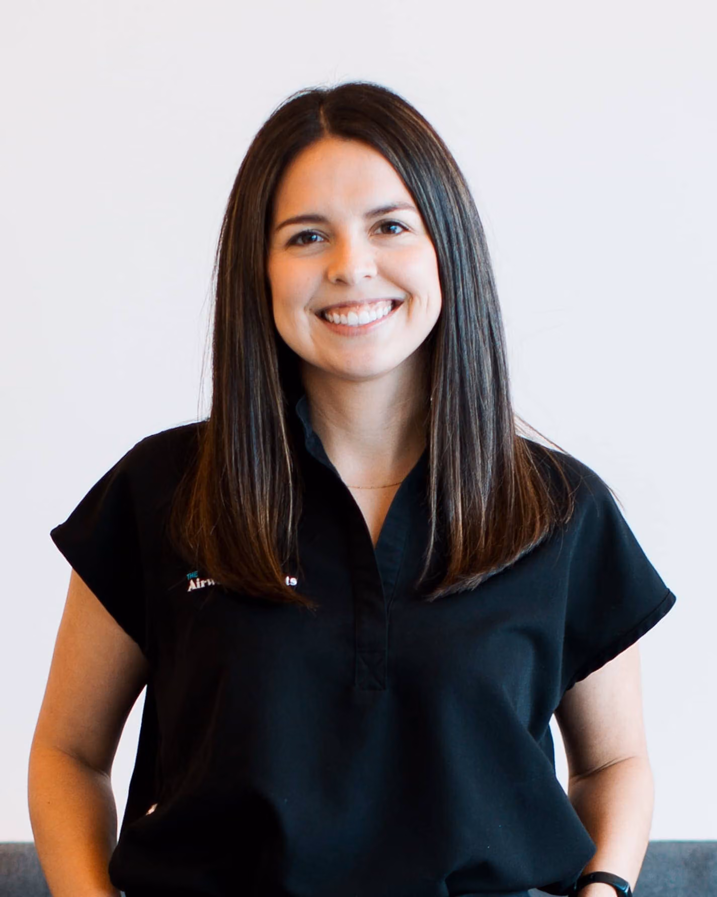 A woman with straight brown hair and a black short-sleeve shirt stands smiling in front of a plain white background.