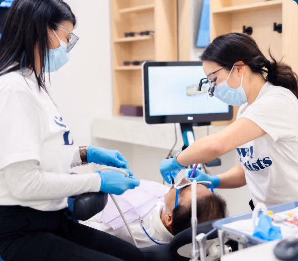 Two clinicians in masks work alongside a patient in a dental chair, with imaging on a monitor in the background.