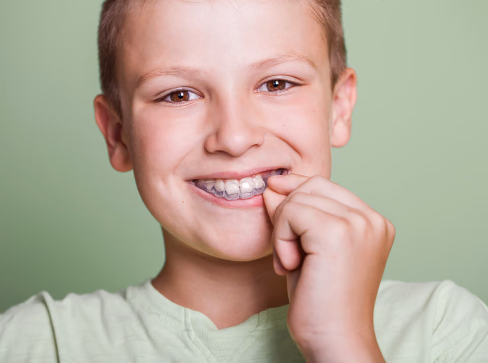 Young boy smiling and placing a clear dental aligner on his upper teeth, wearing a light green shirt against a plain green background.