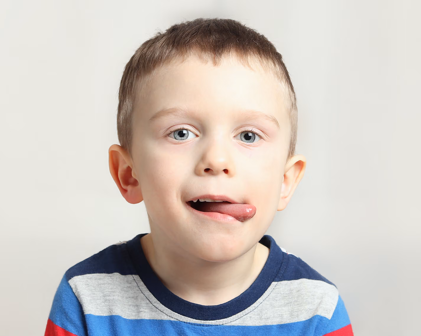 Young boy with short hair wearing a striped shirt sticks out his tongue to the side while looking at the camera, against a plain light background.