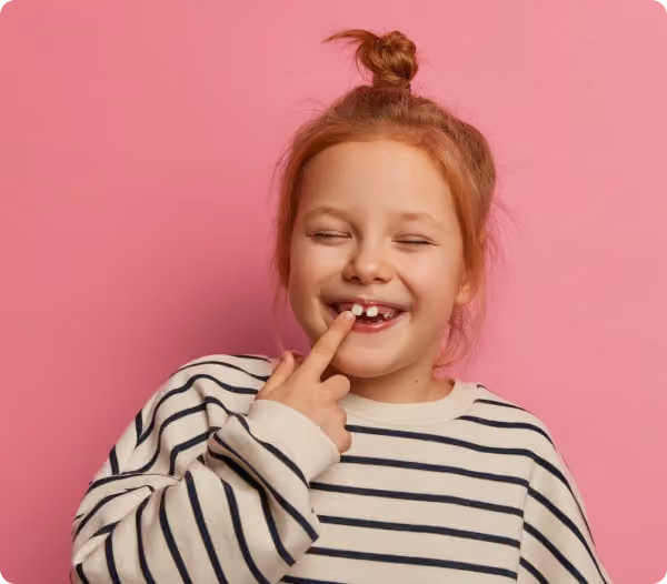 Young girl with red hair in a bun, wearing a striped shirt, smiling and pointing to a missing tooth, standing against a pink background.