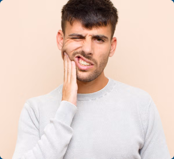 A man in a light gray sweater touches his cheek and winces, suggesting tooth pain, against a beige background.