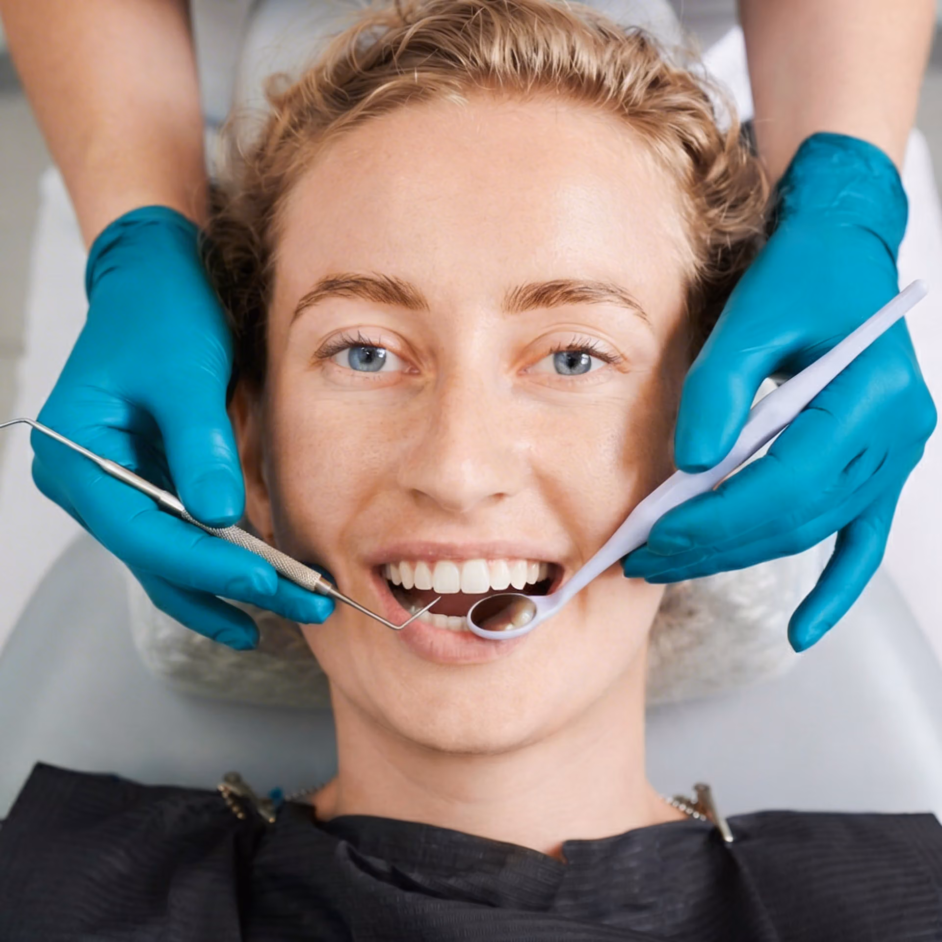 A person sits in a dental chair while a dentist wearing teal gloves examines their teeth with dental instruments.