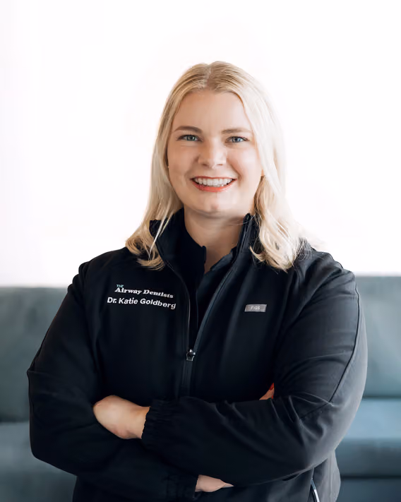 A woman with blonde hair in a black jacket embroidered with “Dr. Katie Goldberg” stands with arms crossed and smiles at the camera.