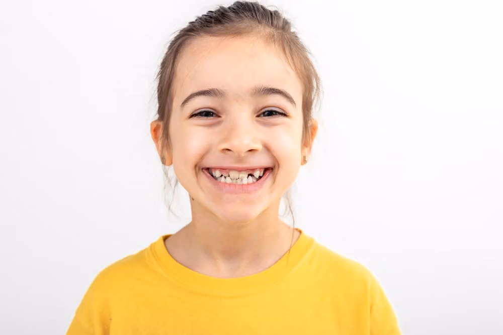 Smiling young girl wearing yellow shirt, missing front teeth visible, showing healthy jaw growth and kids’ smiles against a white background