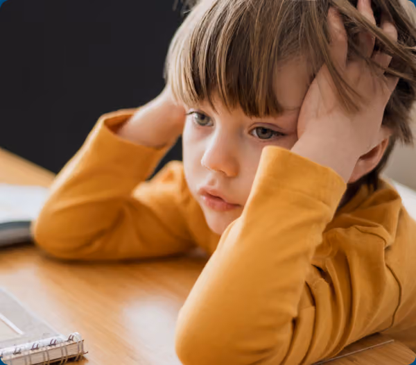 Young boy with hand on his head, looking distracted at a desk — tied to ADHD-like symptoms from poor sleep.