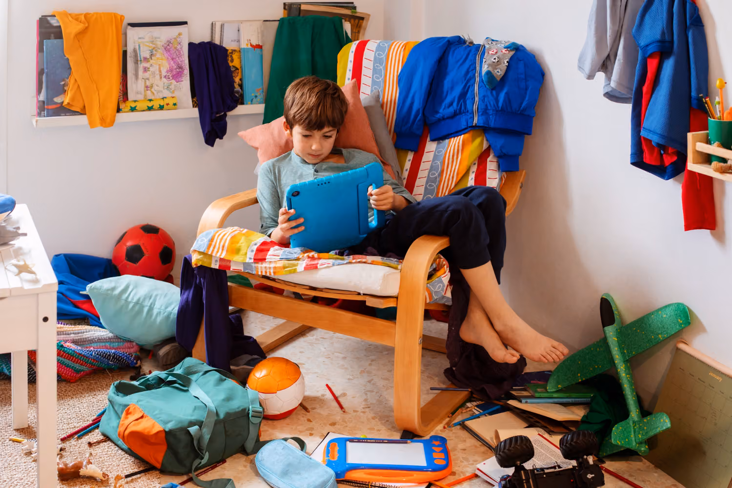 A boy sits barefoot in a colorful, cluttered room on a chair, using a tablet surrounded by scattered toys, clothes, and school supplies.