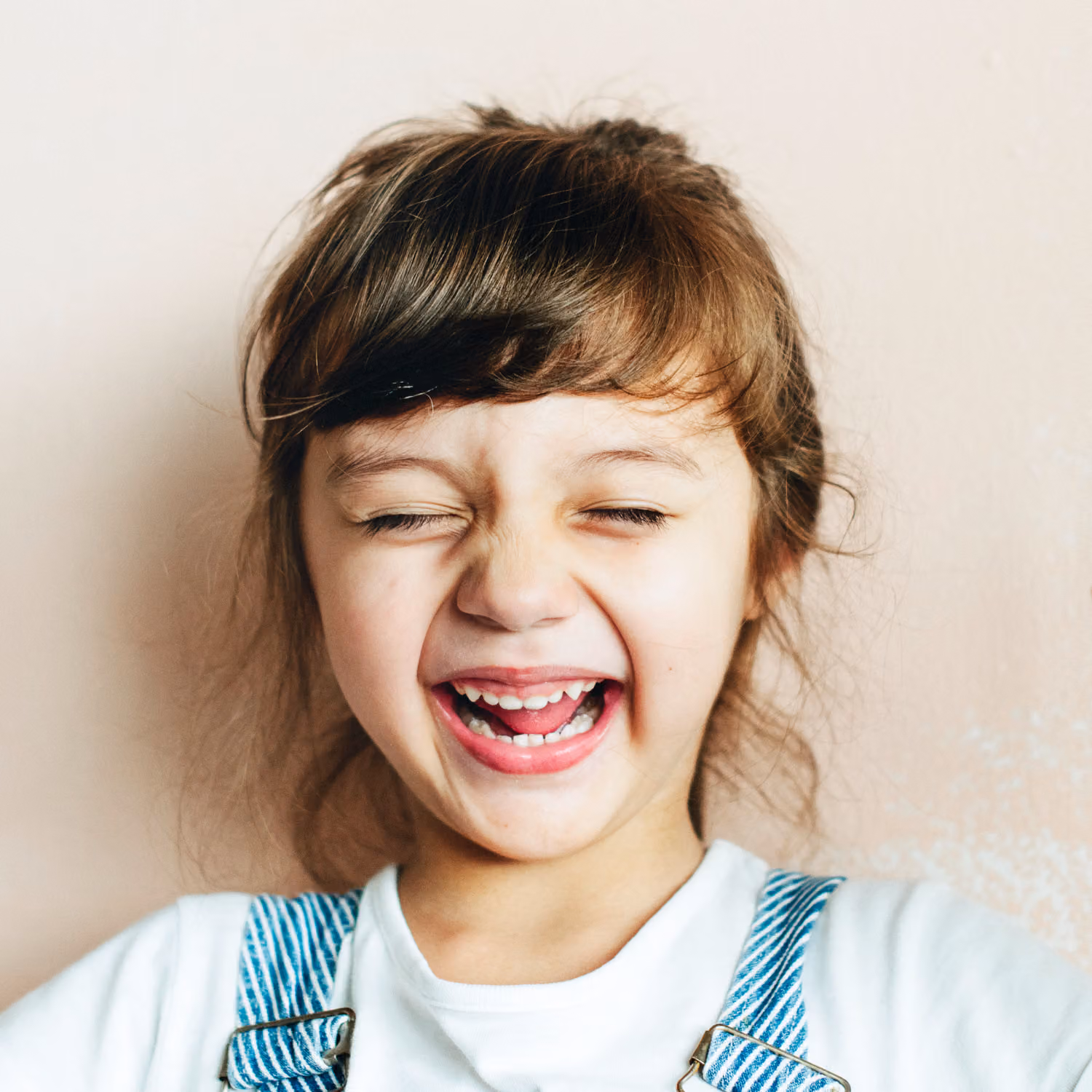 Young child with brown hair wearing a white shirt and denim overalls, smiling widely with eyes closed against a light-colored wall.
