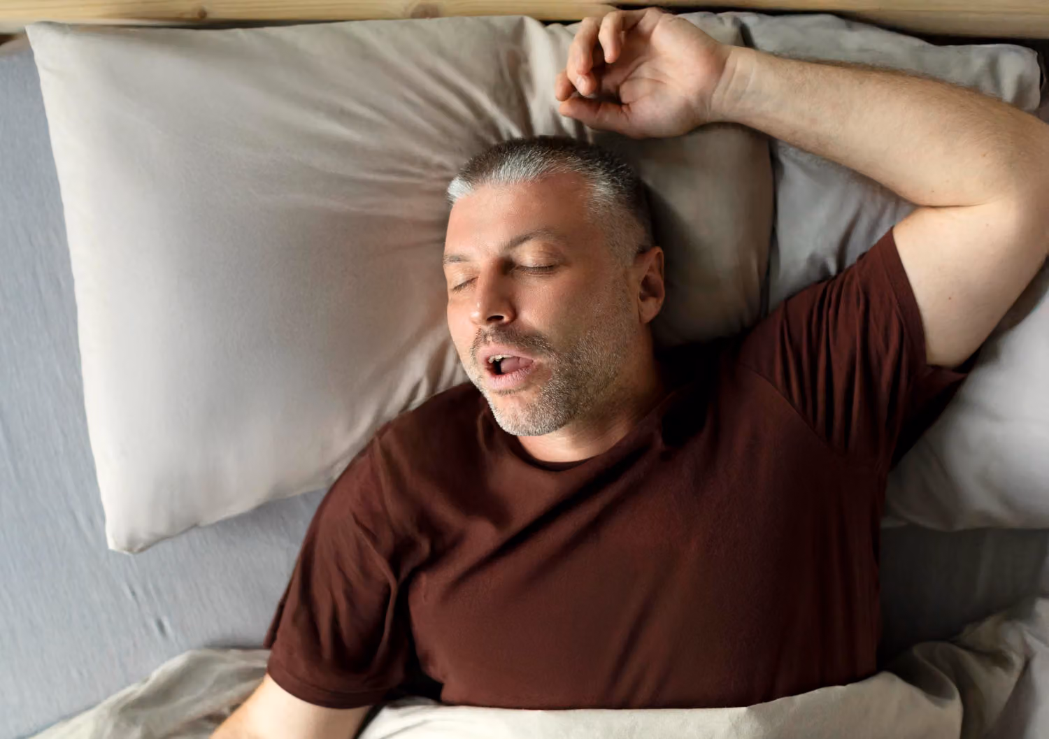A middle-aged man with short gray hair is lying in bed on his back, mouth open, and appears to be sleeping.