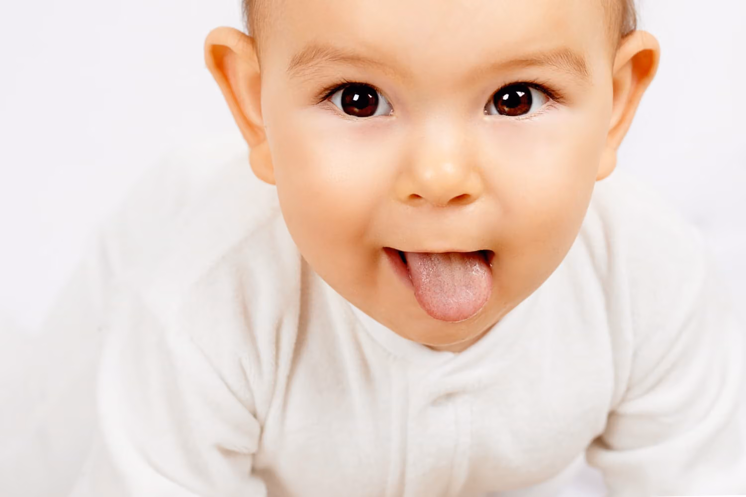 A baby in a white outfit sticks out their tongue, looking up at the camera against a plain light background.