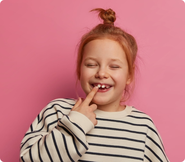 Young girl with red hair in a bun, wearing a striped shirt, smiling and pointing to a missing tooth, standing against a pink background.