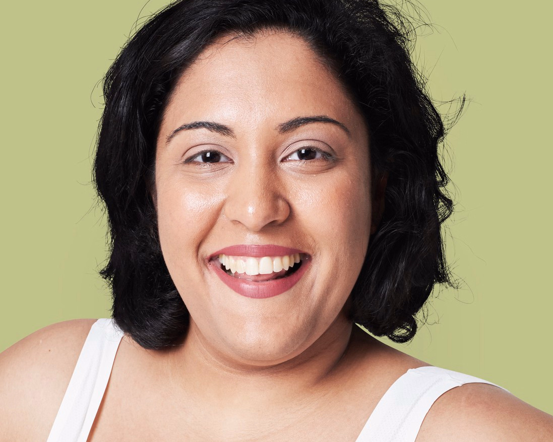 A woman with short black hair and a brown shirt smiles broadly while looking toward the camera against a white background.