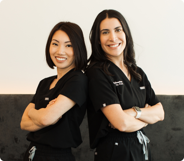 Two women dentists in dark scrubs stand together, smiling with arms crossed.