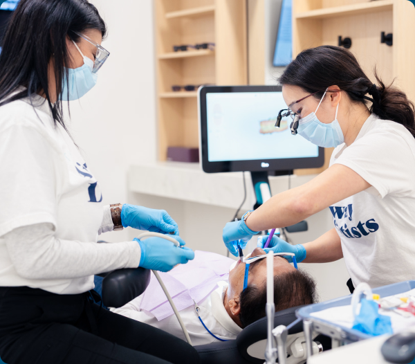 Two clinicians in masks work alongside a patient in a dental chair, with imaging on a monitor in the background.