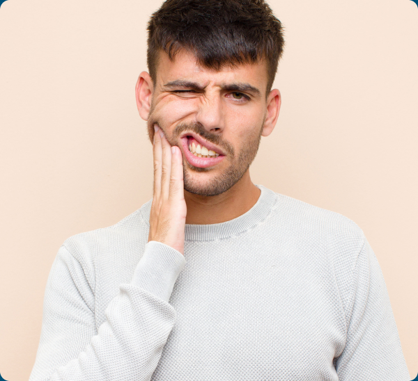A man in a light gray sweater touches his cheek and winces, suggesting tooth pain, against a beige background.