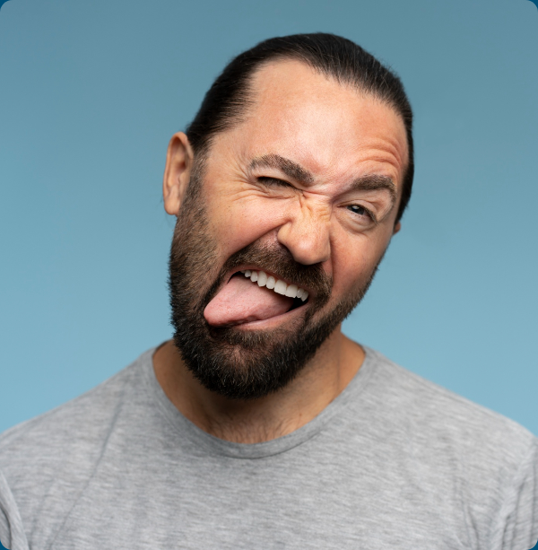 A bearded man sticks out his tongue against a blue studio background.