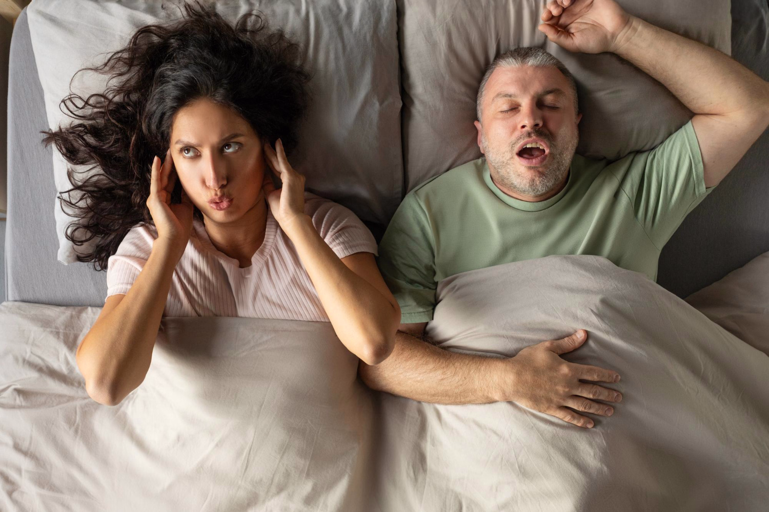 A man snores in bed while his partner covers her ears with her fingers.