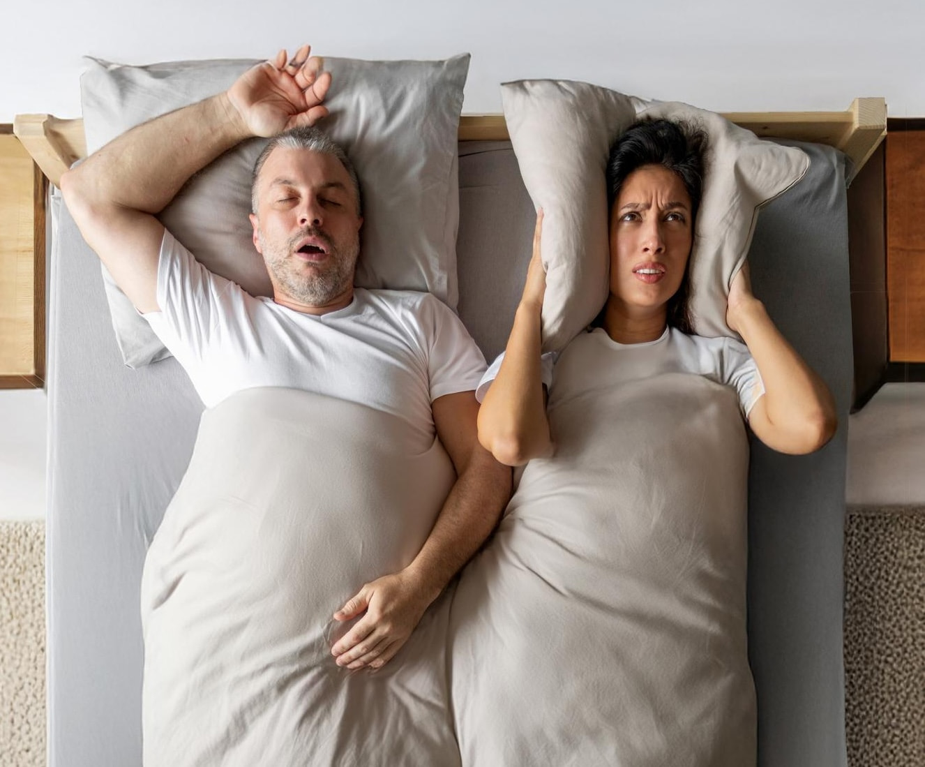 A man is sleeping on his back with his mouth open, while a woman beside him covers her ears and looks annoyed.