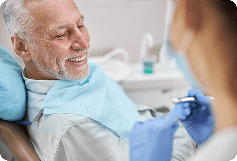 Smiling older man sits in a dental chair while a dentist gets tools ready to discuss dental crown costs in Houston