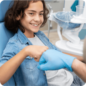 A smiling child sitting in a dental chair gives a fist bump to a person wearing a blue medical glove.