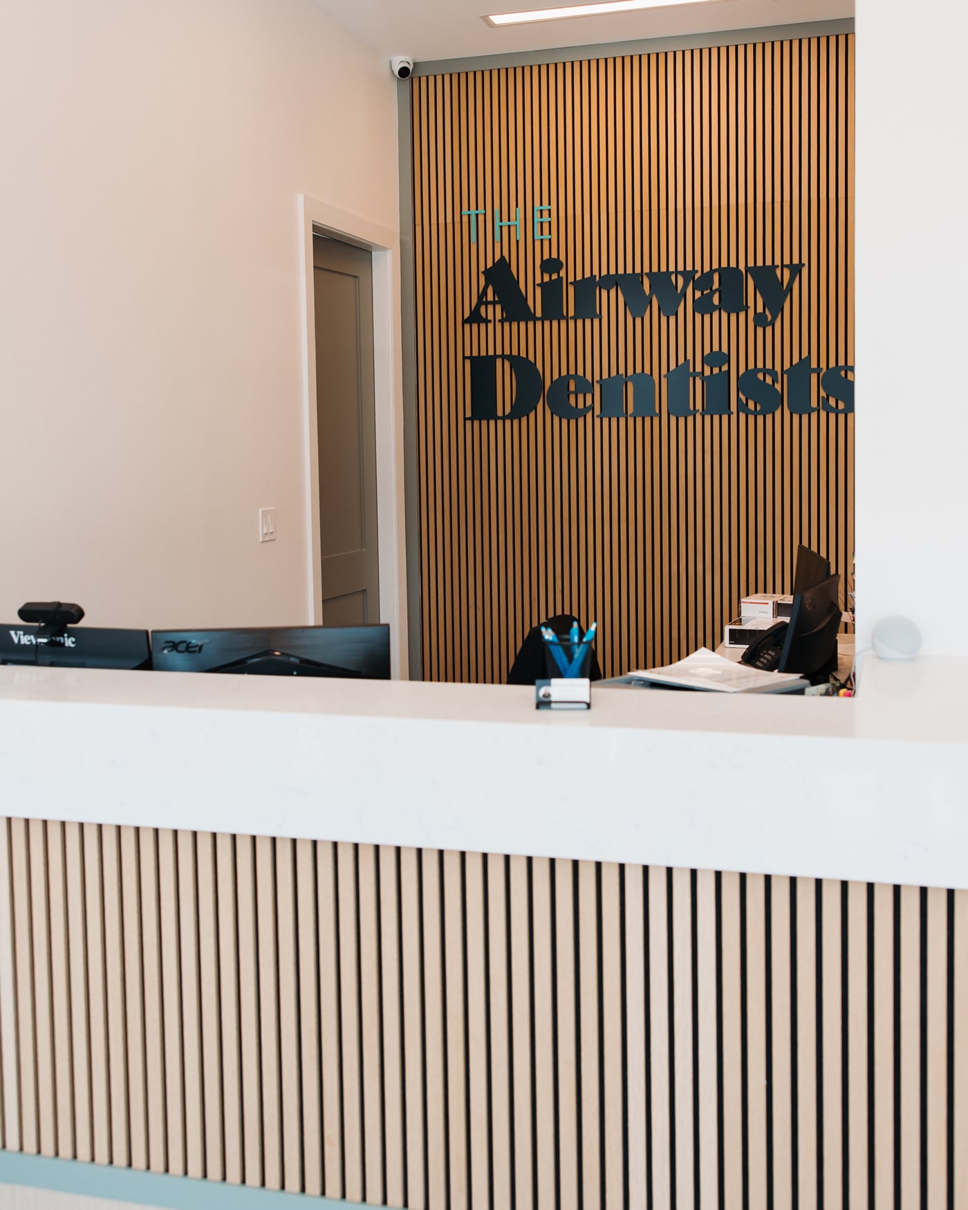 Reception desk with computers and office supplies in front of a wood-paneled wall displaying "The Airway Dentist" signage in a modern dental office.