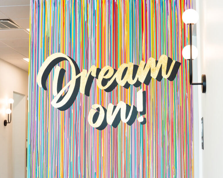 Gold script text reading "Dream on!" is displayed on a colorful wall of hanging ribbons, with round wall lights visible on either side.