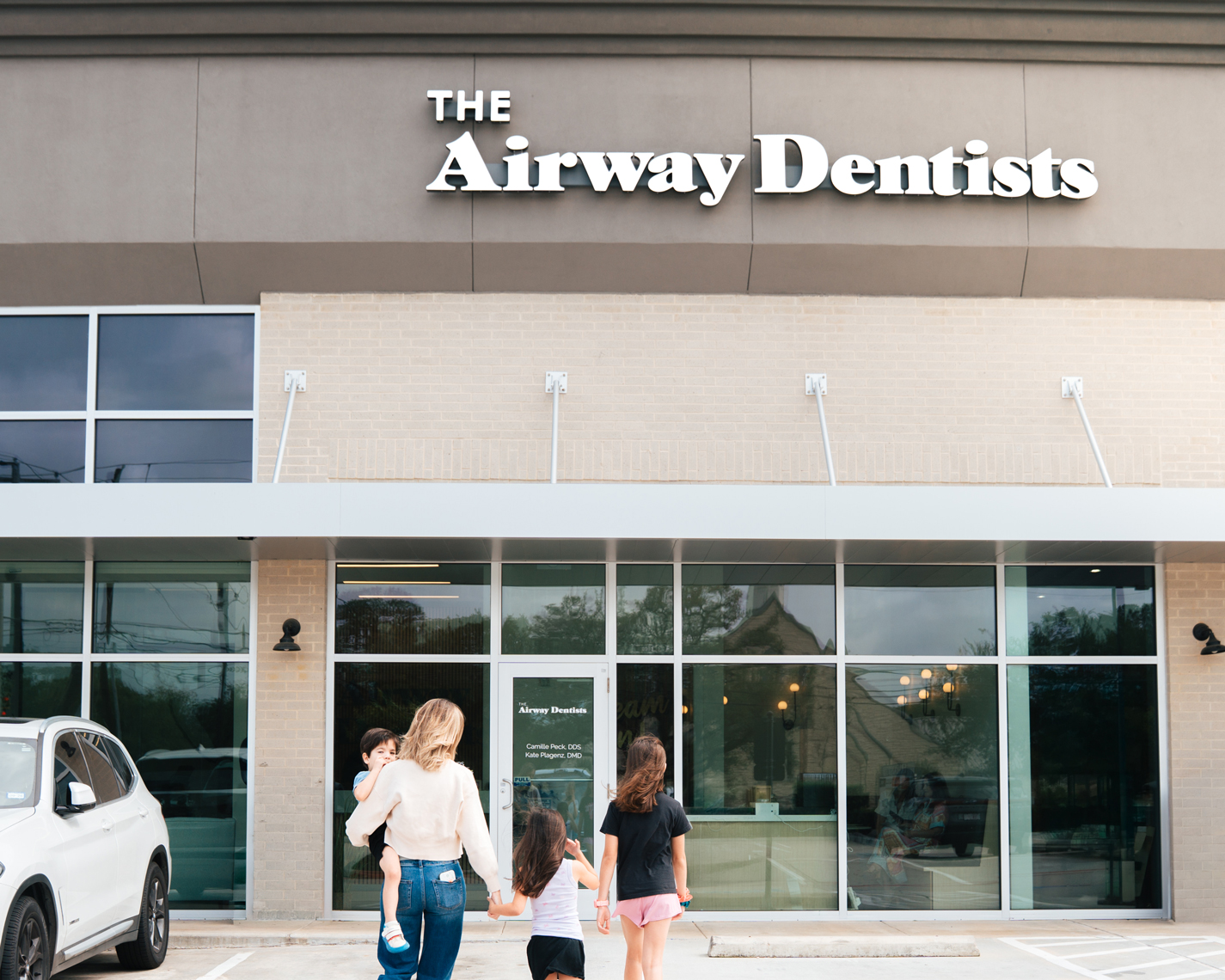 A woman and three children walk toward the entrance of a dental office called "The Airway Dentists" in a modern building with large glass doors.