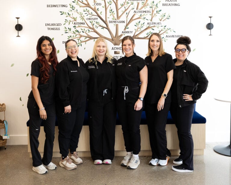Six women in black medical scrubs stand and smile in front of a wall with a tree graphic listing various health conditions.