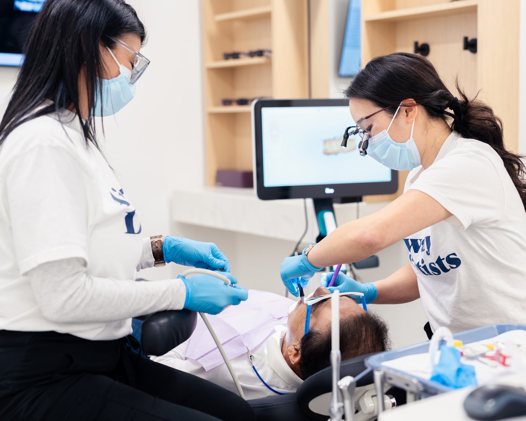 Two dental professionals in masks and gloves perform a dental procedure on a patient in a modern clinic setting with equipment visible.