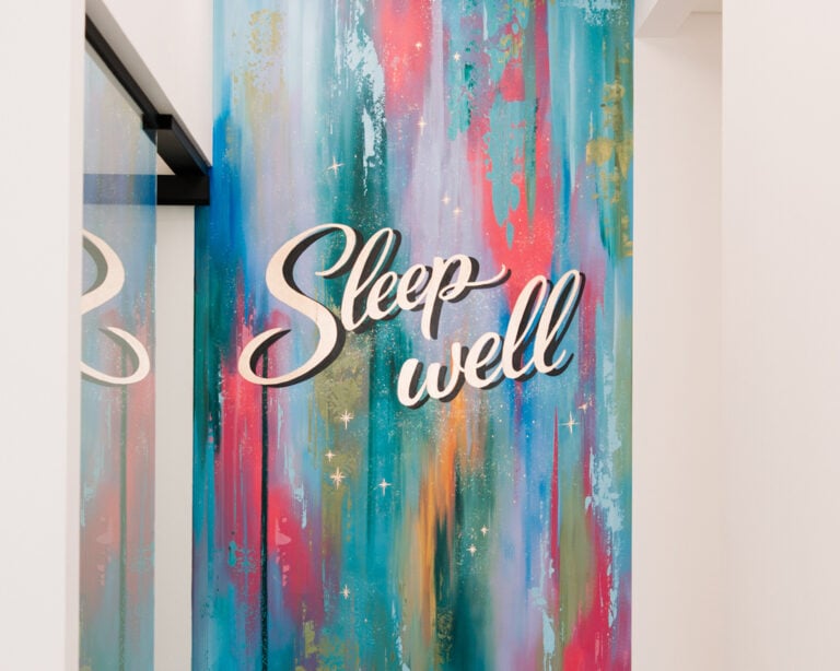 A colorful wall mural features the phrase "Sleep well" in cursive text against a vibrant, abstract painted background.