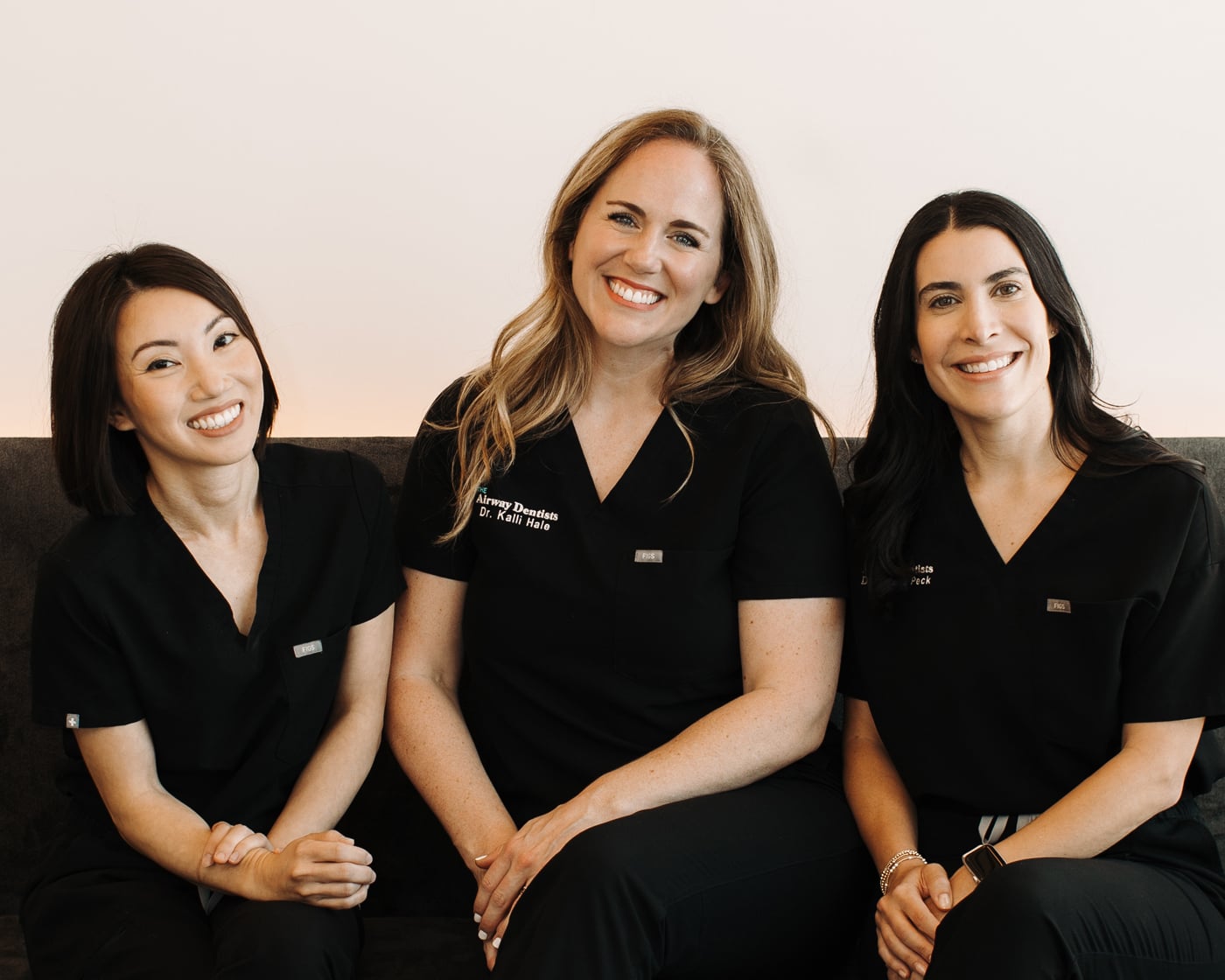 Three members of The Airway Dentists sitting and smiling wearing matching black "Airway Dentists" scrubs.