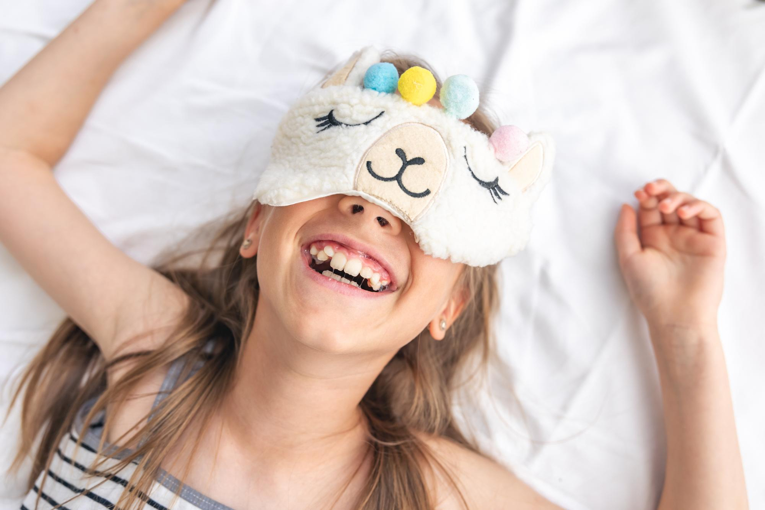 Smiling child lies on a bed wearing a sleep mask with a cartoon animal face and colorful pom-poms, dressed in a striped tank top.