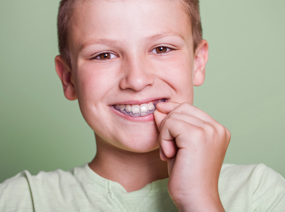 Young boy smiling and placing a clear dental aligner on his upper teeth, wearing a light green shirt against a plain green background.