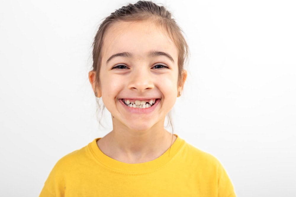 Smiling young girl wearing yellow shirt, missing front teeth visible, showing healthy jaw growth and kids’ smiles against a white background