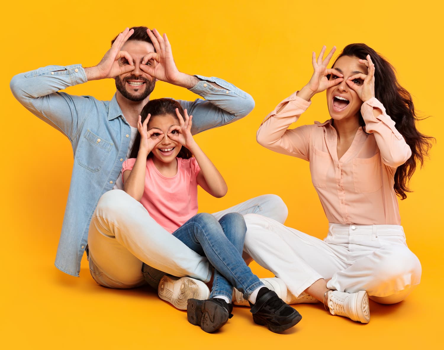 Father, mother, and daughter sitting together on a yellow backdrop, making glasses on their faces with their hands