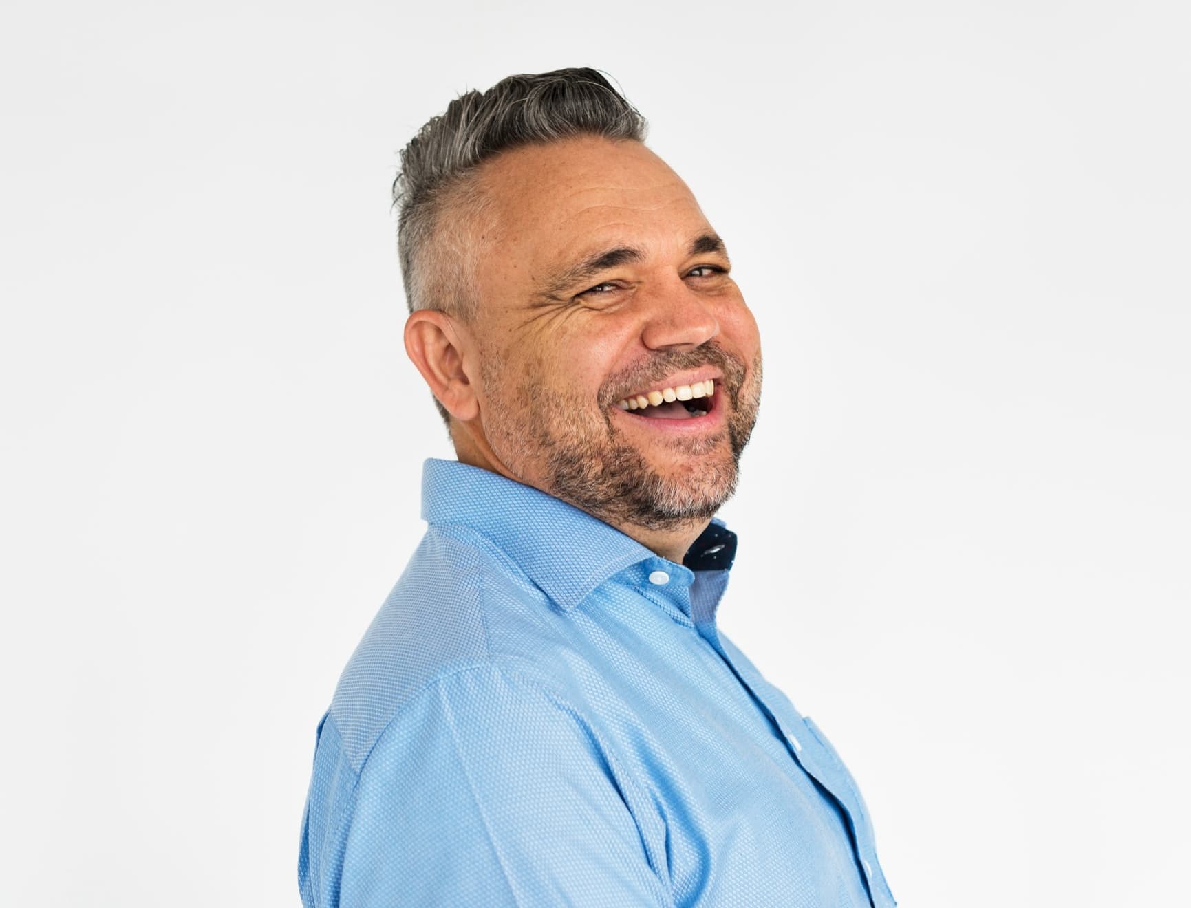 Middle-aged man with short gray hair and a beard, wearing a light blue shirt, smiling and looking to the side against a plain white background.