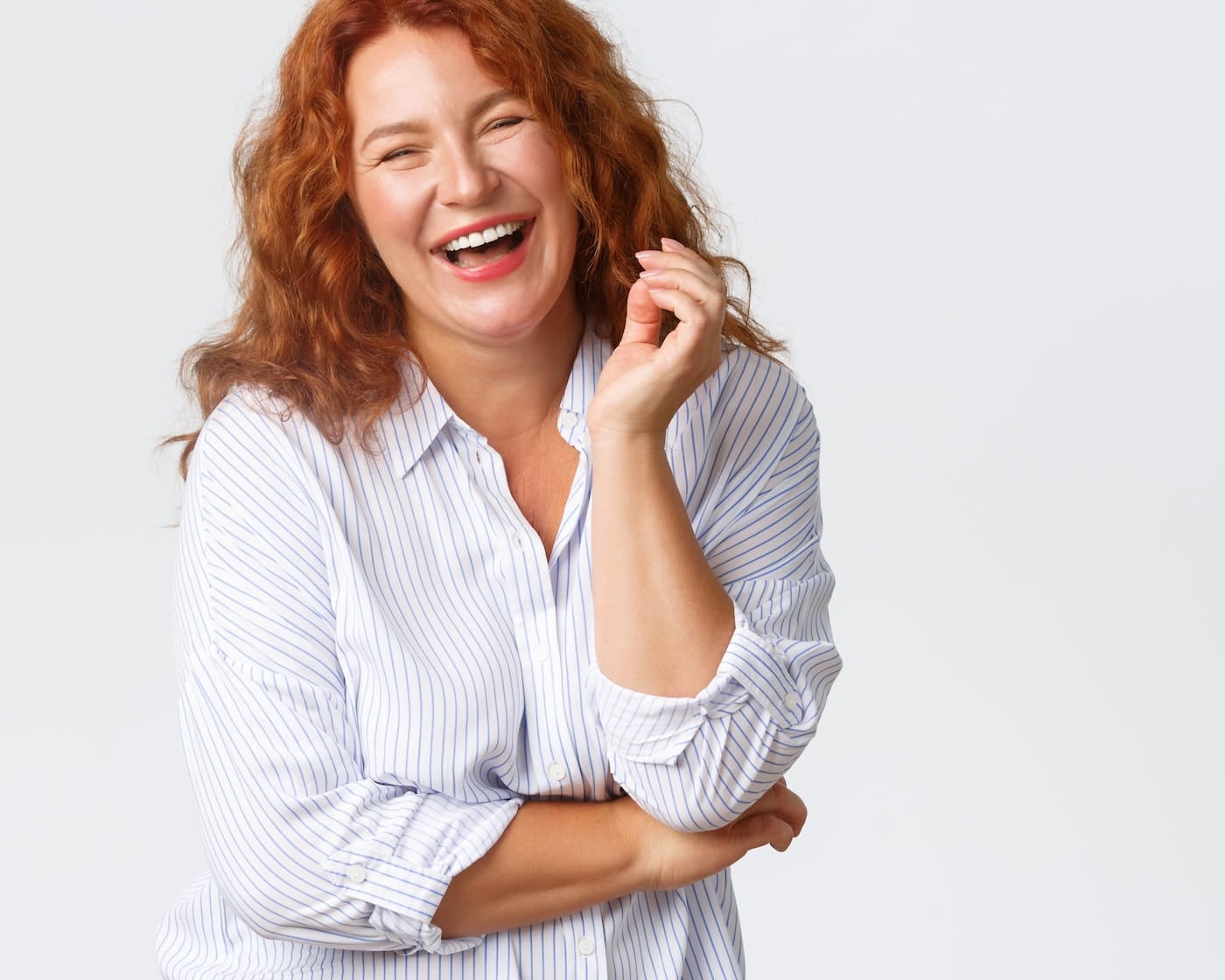 Woman with long, curly red hair wearing a striped shirt stands against a plain background, smiling and laughing with one arm crossed and the other hand raised.