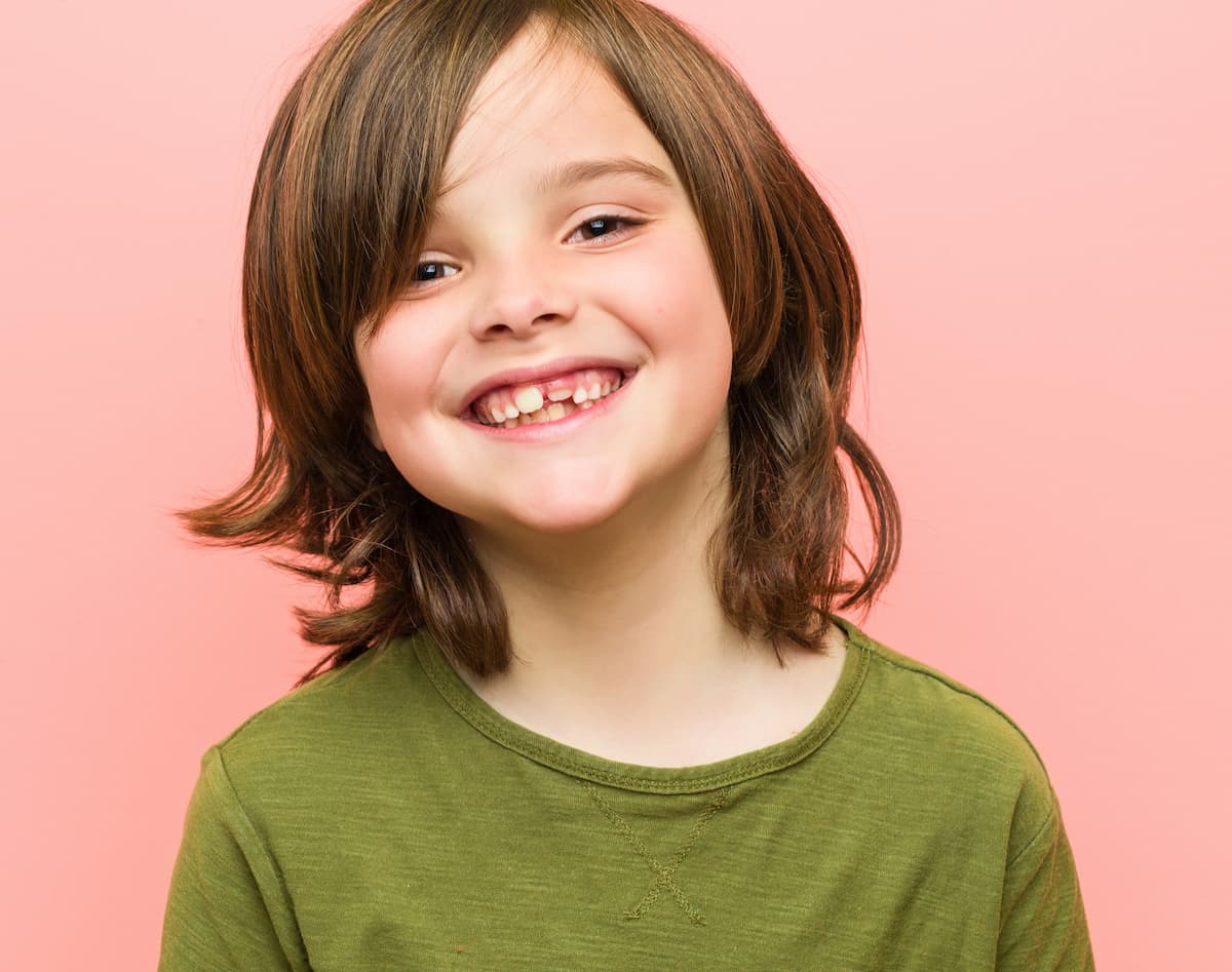 Smiling boy with a missing front tooth in a green shirt against a pink background