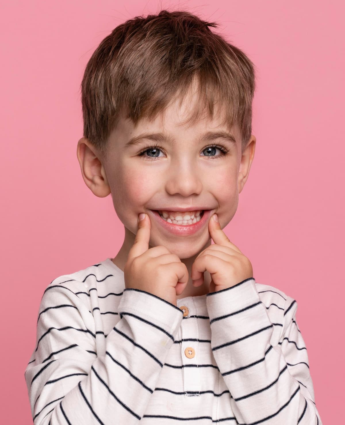 Smiling young boy in a white pinstriped shirt, pointing at his smile against a pink background