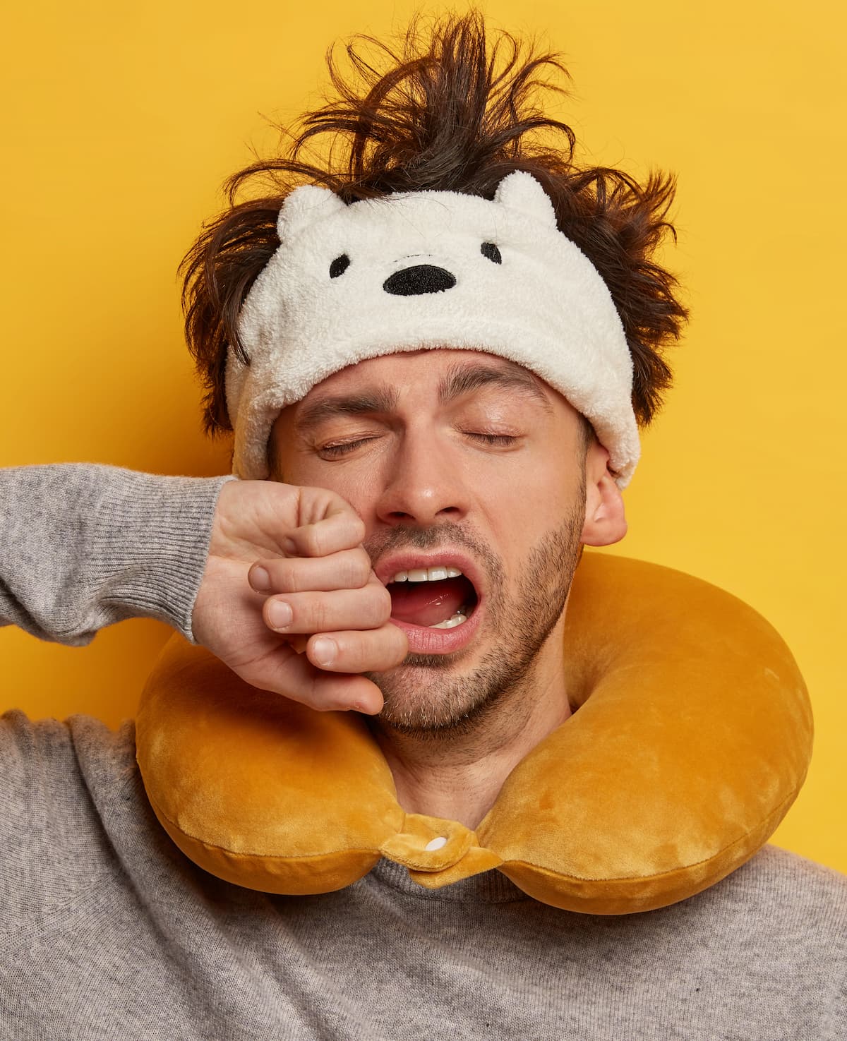 Sleepy man with frazzled hair, wearing a fuzzy bear sleep mask and neck pillow, yawning against a yellow background