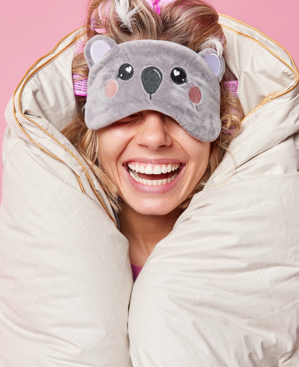 Smiling woman wearing a koala sleep mask, cuddled in a blanket against a pink background